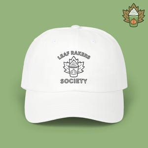 May include: A white baseball cap featuring a black graphic of a maple leaf behind a cup with whipped cream and a small pumpkin icon. The text "LEAF RAKERS SOCIETY" is printed in black capital letters above and below the central graphic design.