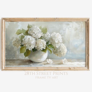 Op de afbeelding: Een ingelijst schilderij van een boeket witte hortensia's in een witte vaas op een houten tafel. De achtergrond is een zachte blauwe en witte kleurverloop.