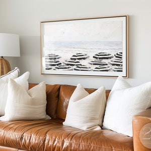 K&ouml;nnte beinhalten: Ein braunes Ledersofa mit wei&szlig;en Kissen. &Uuml;ber dem Sofa h&auml;ngt ein gerahmtes Schwarzwei&szlig;bild mit einer Strandszene.