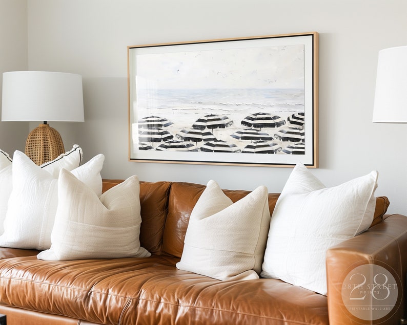 K&ouml;nnte beinhalten: Ein braunes Ledersofa mit wei&szlig;en Kissen. &Uuml;ber dem Sofa h&auml;ngt ein gerahmtes Schwarzwei&szlig;bild mit einer Strandszene.