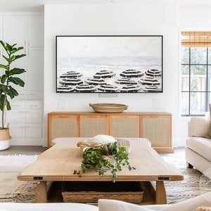 K&ouml;nnte beinhalten: Ein Wohnzimmer-Interieur mit einem Couchtisch aus hellem Holz, einem wei&szlig;en Sofa und einem gro&szlig;en, schwarz gerahmten Gem&auml;lde einer Strandszene mit schwarz-wei&szlig;en Sonnenschirmen. In der Ecke des Raumes steht eine gro&szlig;e Topfpflanze.