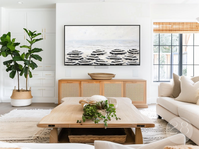 K&ouml;nnte beinhalten: Ein Wohnzimmer-Interieur mit einem Couchtisch aus hellem Holz, einem wei&szlig;en Sofa und einem gro&szlig;en, schwarz gerahmten Gem&auml;lde einer Strandszene mit schwarz-wei&szlig;en Sonnenschirmen. In der Ecke des Raumes steht eine gro&szlig;e Topfpflanze.