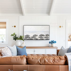 K&ouml;nnte beinhalten: Ein gro&szlig;es braunes Ledersofa in L-Form mit verschiedenen Kissen in Beige-, Blau- und Wei&szlig;t&ouml;nen. Das Sofa steht vor einer wei&szlig;en Wand mit einem gro&szlig;en abstrakten Gem&auml;lde dar&uuml;ber. Das Gem&auml;lde zeigt ein blaues und wei&szlig;es Farbschema und stellt eine Strandszene dar.