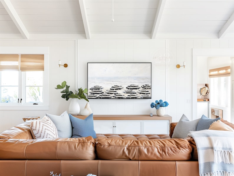 K&ouml;nnte beinhalten: Ein gro&szlig;es braunes Ledersofa in L-Form mit verschiedenen Kissen in Beige-, Blau- und Wei&szlig;t&ouml;nen. Das Sofa steht vor einer wei&szlig;en Wand mit einem gro&szlig;en abstrakten Gem&auml;lde dar&uuml;ber. Das Gem&auml;lde zeigt ein blaues und wei&szlig;es Farbschema und stellt eine Strandszene dar.