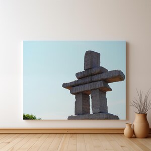 May include: A rectangular wall art print featuring a stone inukshuk structure against a pale blue sky. The sculpture is composed of stacked, rough-hewn grey stone blocks arranged in a human-like form, positioned centrally within the frame.