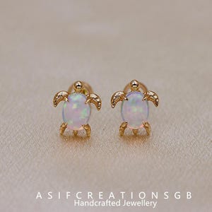 May include: A pair of gold-colored turtle stud earrings. Each earring features an oval, iridescent opal stone set within a turtle-shaped frame. The earrings are detailed with tiny legs, flippers, and a head. The text "ASIF CREATIONS SGB Handcrafted Jewellery" is at the bottom.