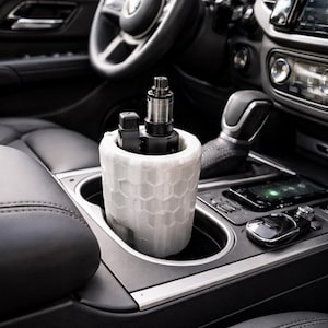 May include: A white, honeycomb-patterned cup holder for a car's center console, holding a vape device and a small black accessory. The interior of a car is visible, with a steering wheel and dashboard in the background.