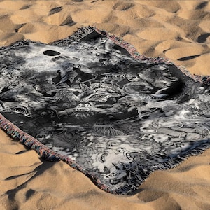 May include: A black and white woven blanket with a detailed graphic design featuring various creatures and abstract shapes. The blanket has a fringed edge with red, blue, and yellow threads. The blanket is displayed on a sandy surface.