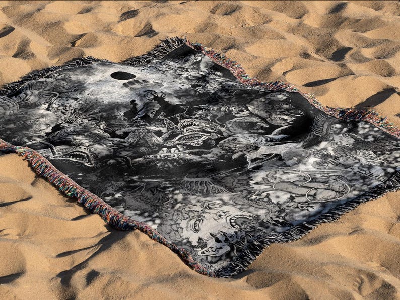 May include: A black and white woven blanket with a detailed graphic design featuring various creatures and abstract shapes. The blanket has a fringed edge with red, blue, and yellow threads. The blanket is displayed on a sandy surface.