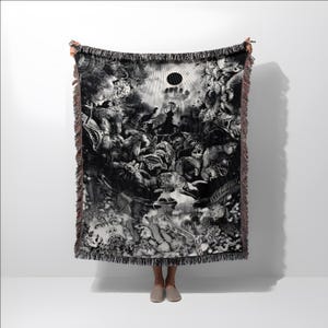 May include: Black and white woven throw blanket featuring a detailed, surreal illustration. The artwork depicts a chaotic scene with figures, a central sun-like shape, and fringed edges. The blanket is held up against a white background.