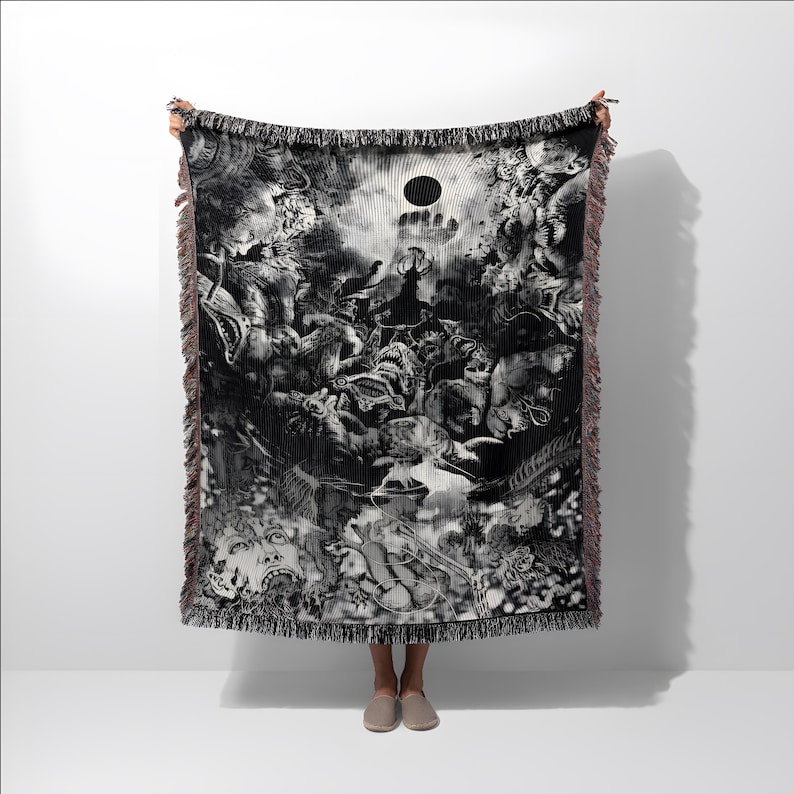 May include: Black and white woven throw blanket featuring a detailed, surreal illustration. The artwork depicts a chaotic scene with figures, a central sun-like shape, and fringed edges. The blanket is held up against a white background.