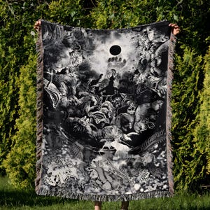 May include: Black and white woven blanket with a detailed, fantastical illustration. The design features a central dark circle, surrounded by intricate figures and creatures. The blanket has fringed edges and is held up in front of a green background.
