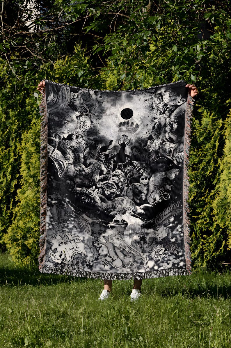 May include: Black and white woven blanket with a detailed, fantastical illustration. The design features a central dark circle, surrounded by intricate figures and creatures. The blanket has fringed edges and is held up in front of a green background.