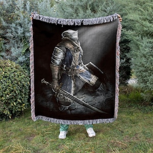 May include: A black woven throw blanket featuring a detailed illustration of a knight in armor, holding a sword. The knight is kneeling on a stone surface. The blanket has a fringed edge.