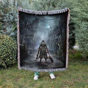 Puede incluir: Una manta tejida que representa una escena oscura y gótica con una figura con abrigo largo sosteniendo armas, sobre un paisaje urbano detallado. La manta tiene un borde con flecos y un diseño detallado, ideal para fans de fantasía o videojuegos.