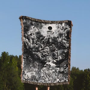 May include: A black and white woven throw blanket with a detailed, surreal design. The blanket has a fringed edge and a circular cutout. The image is taken outdoors, with a blue sky and green trees in the background.