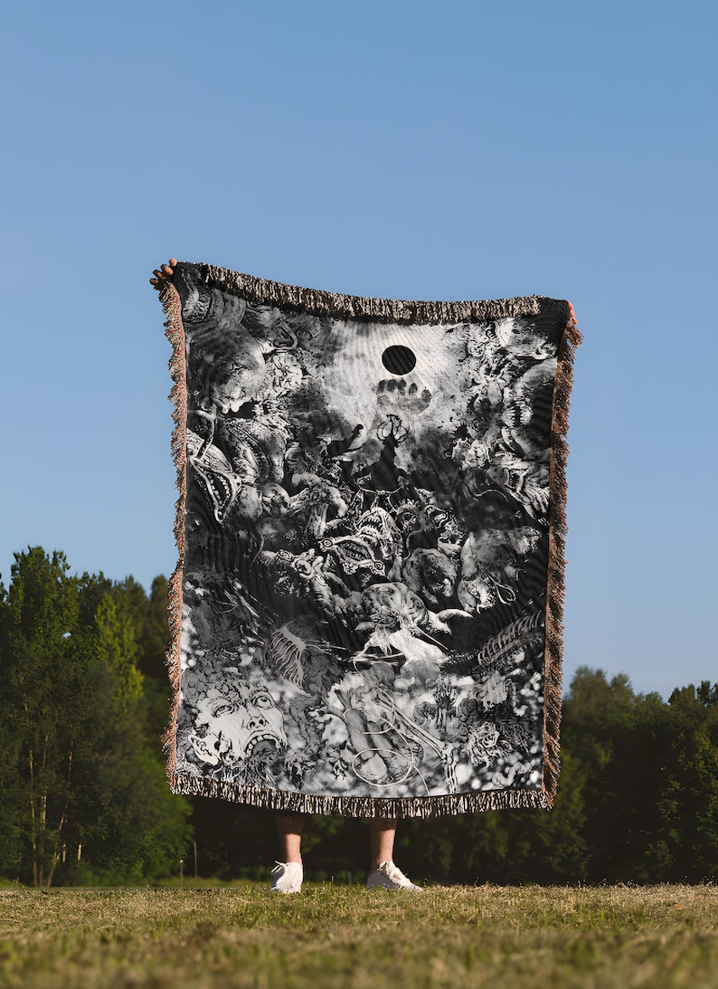May include: A black and white woven throw blanket with a detailed, surreal design. The blanket has a fringed edge and a circular cutout. The image is taken outdoors, with a blue sky and green trees in the background.