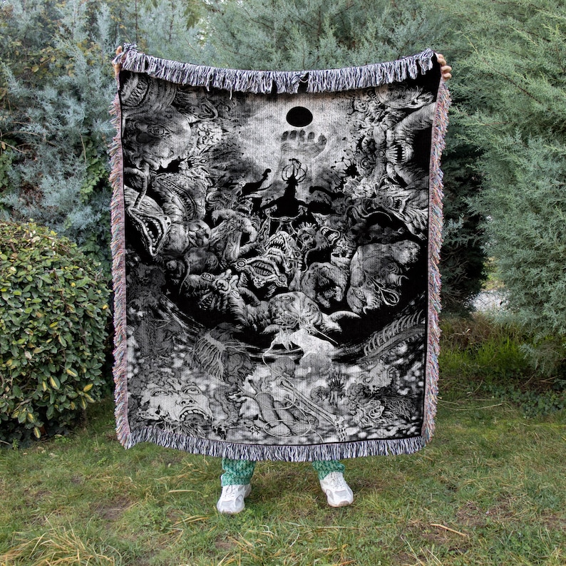 May include: Black and white woven throw blanket featuring a detailed illustration of fantastical creatures and a central sun-like design. The blanket has a fringed edge and is held up against a backdrop of green foliage.