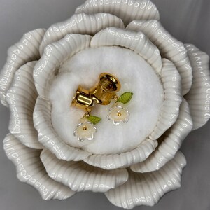 K&ouml;nnte beinhalten: Ein Paar goldene Ohrdehner mit wei&szlig;en Blumenakzenten. Die Dehnungen werden in einer wei&szlig;en, rosenf&ouml;rmigen Keramikschale pr&auml;sentiert.