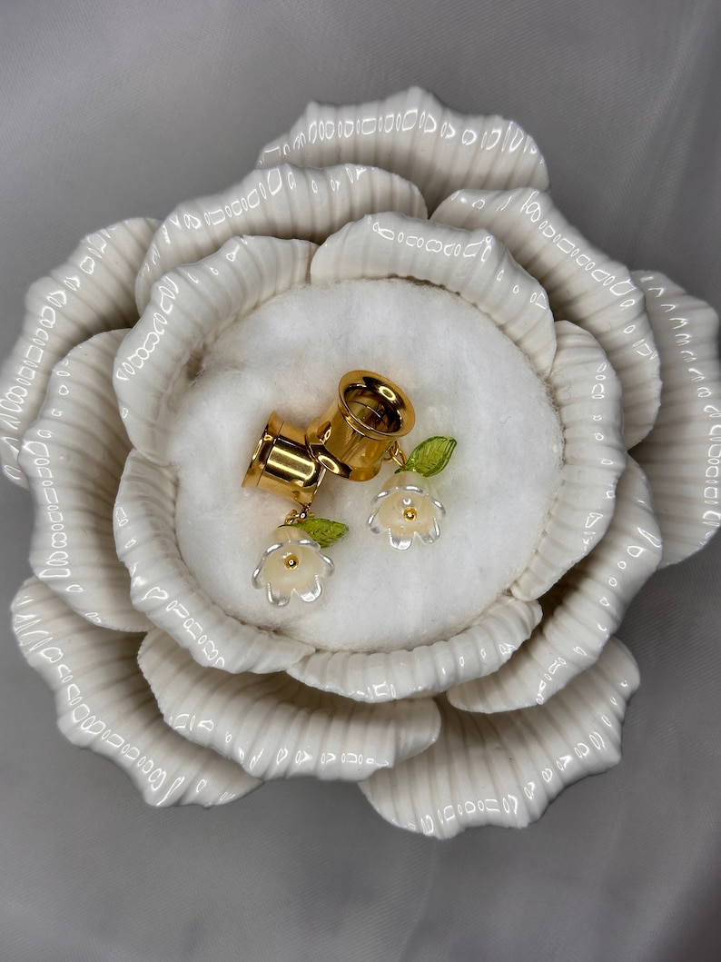 K&ouml;nnte beinhalten: Ein Paar goldene Ohrdehner mit wei&szlig;en Blumenakzenten. Die Dehnungen werden in einer wei&szlig;en, rosenf&ouml;rmigen Keramikschale pr&auml;sentiert.