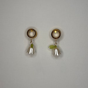 K&ouml;nnte beinhalten: Ein Paar goldfarbene Ohrdehner mit wei&szlig;en Bl&uuml;tenanh&auml;ngern. Die Dehnungen sind rund und haben eine glatte Oberfl&auml;che. Die Bl&uuml;ten haben gr&uuml;ne Bl&auml;tter.