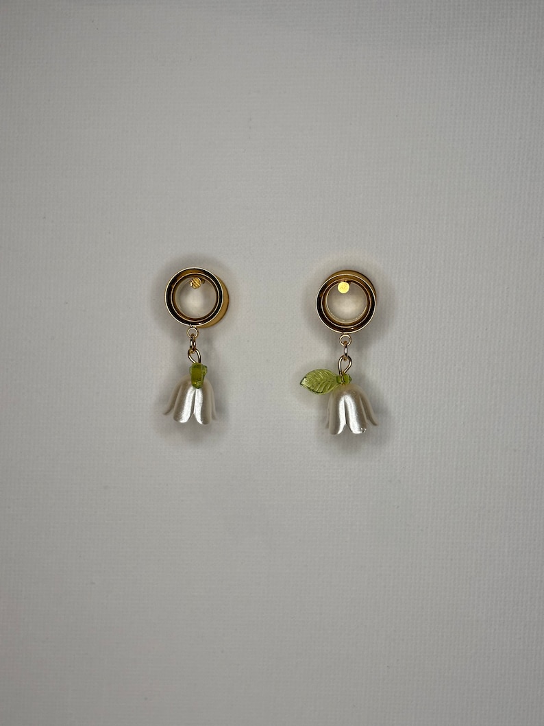 K&ouml;nnte beinhalten: Ein Paar goldfarbene Ohrdehner mit wei&szlig;en Bl&uuml;tenanh&auml;ngern. Die Dehnungen sind rund und haben eine glatte Oberfl&auml;che. Die Bl&uuml;ten haben gr&uuml;ne Bl&auml;tter.