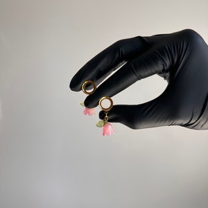 K&ouml;nnte beinhalten: Ein Paar goldene Creolen mit rosa Blumenanh&auml;ngern. Die Ohrringe werden in einer behandschuhten Hand gehalten.
