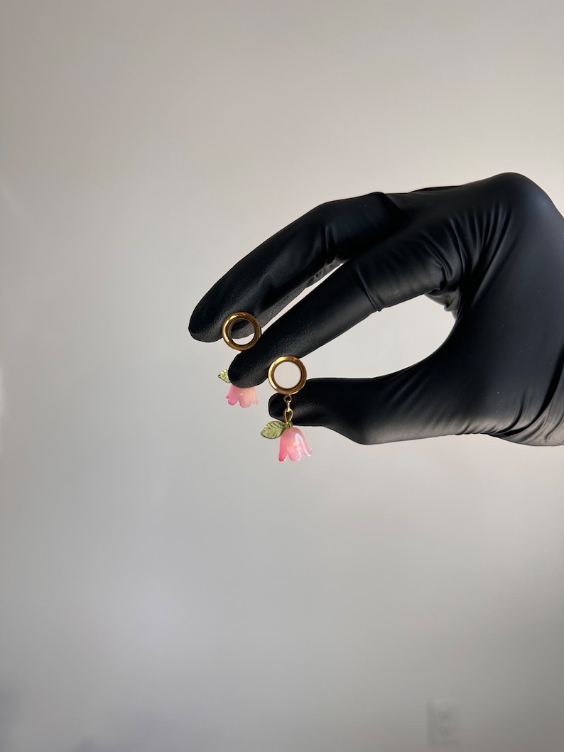 K&ouml;nnte beinhalten: Ein Paar goldene Creolen mit rosa Blumenanh&auml;ngern. Die Ohrringe werden in einer behandschuhten Hand gehalten.