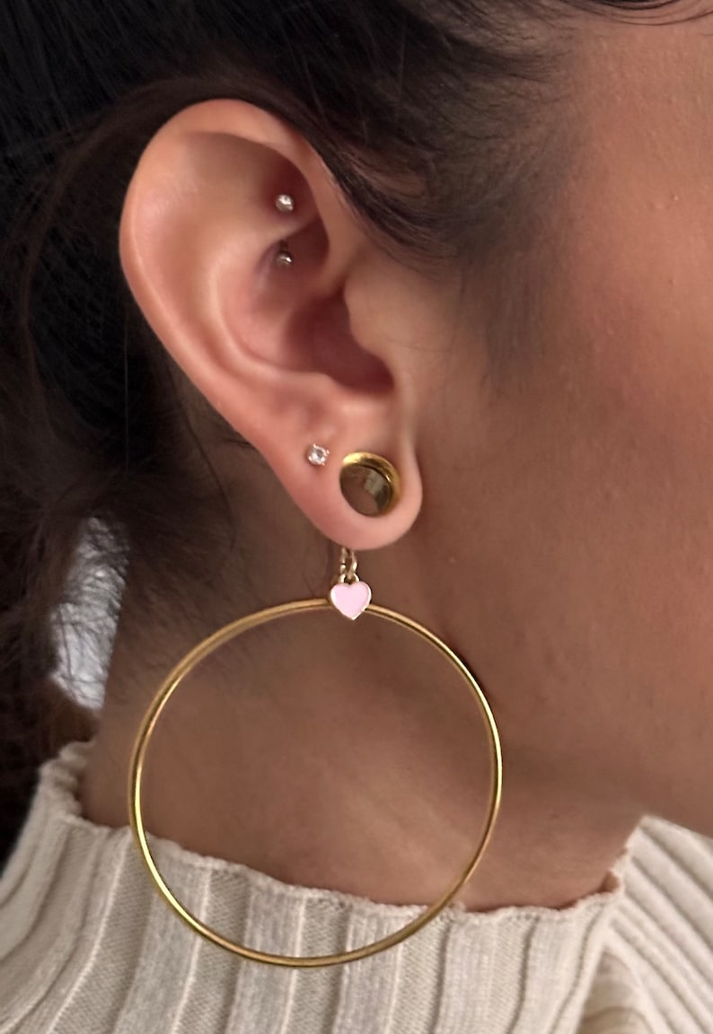 May include: Gold hoop earrings with a small pink heart charm hanging from one earring. The earrings are worn in a person's ear with a gold ear gauge and two small diamond studs.