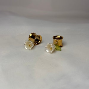 May include: A pair of gold ear gauges with white flower charms. The gauges are made of metal and have a smooth finish. The flowers are delicate and have a realistic appearance.