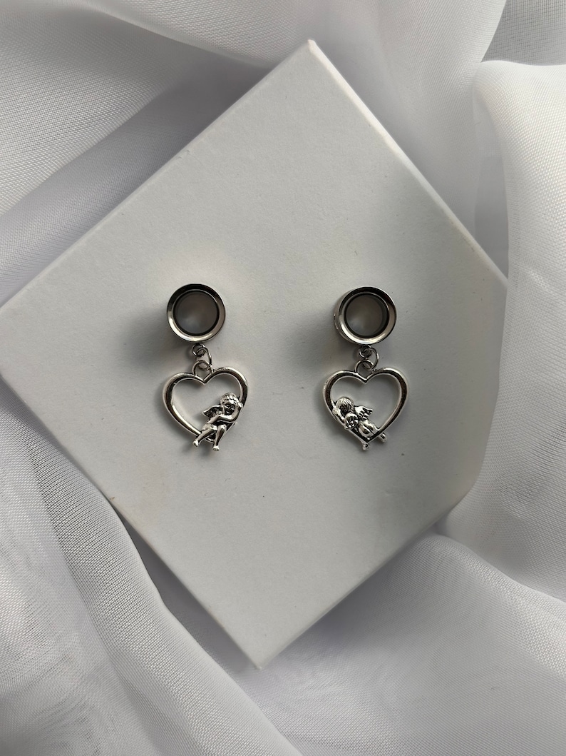 Puede incluir: Un par de pendientes de plata con un dise&ntilde;o en forma de coraz&oacute;n que presenta un querub&iacute;n en su interior. Los pendientes tienen una base redonda de plata.