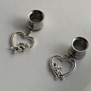 Puede incluir: Un par de dilatadores de oreja plateados con un colgante de coraz&oacute;n que presenta un querub&iacute;n. El colgante de coraz&oacute;n est&aacute; unido al dilatador con una peque&ntilde;a cadena.