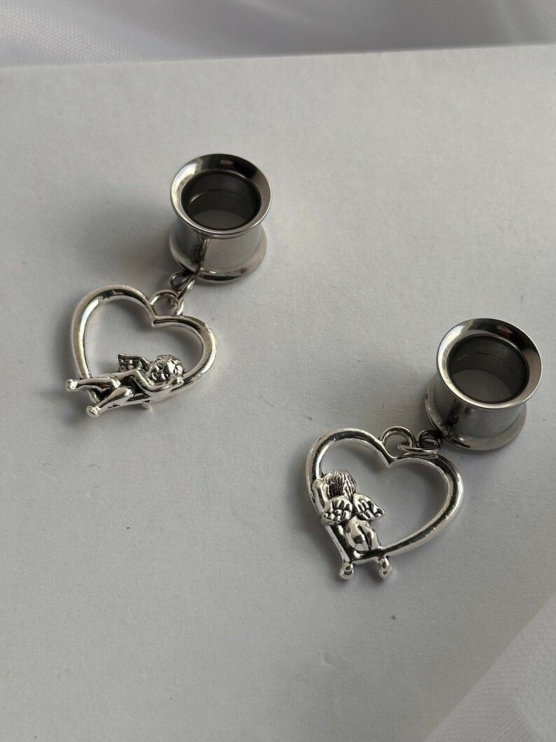 Puede incluir: Un par de dilatadores de oreja plateados con un colgante de coraz&oacute;n que presenta un querub&iacute;n. El colgante de coraz&oacute;n est&aacute; unido al dilatador con una peque&ntilde;a cadena.