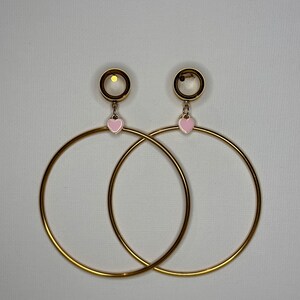 May include: A pair of gold hoop earrings with a pink heart charm dangling from each hoop. The hoops are thick and have a shiny finish.