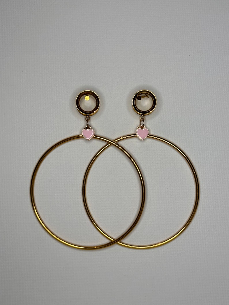 May include: A pair of gold hoop earrings with a pink heart charm dangling from each hoop. The hoops are thick and have a shiny finish.