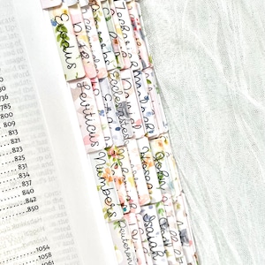 May include: A Bible with floral-patterned tabs marking the books of the Bible. The tabs are white with a watercolor floral design and handwritten book names in black ink. The book is open to a page with numbers.