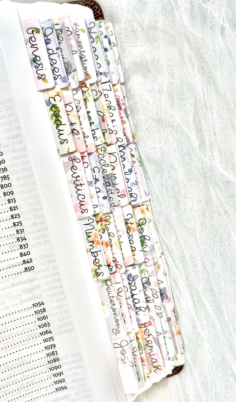 Puede incluir: Un conjunto de marcadores de libros con estampado floral de colores con los nombres de los libros de la Biblia escritos en cursiva negra. Los marcadores est&aacute;n dispuestos en una pila, con los nombres de los libros visibles. Los marcadores son una excelente manera de llevar un registro de tu lugar en la Biblia.