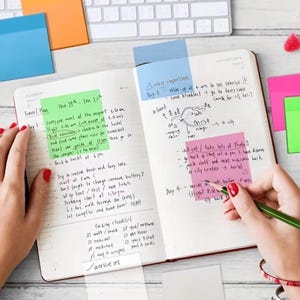 May include: Open notebook with handwritten travel plans, colorful sticky notes, and a keyboard in the background. A hand with red nail polish is writing with a green pen. The notebook is open to two pages with notes and a travel checklist.