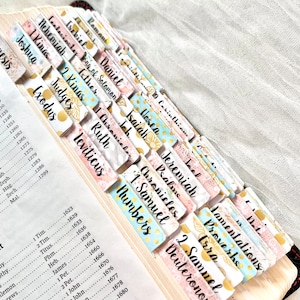 May include: A close-up of a Bible with colorful tabs marking different books of the Bible. The tabs are decorated with floral patterns and gold polka dots. The tabs are labeled with the names of the books of the Bible, including Genesis, Exodus, Leviticus, Numbers, Deuteronomy, Joshua, Judges, Ruth, 1 Samuel, 2 Samuel, 1 Kings, 2 Kings, 1 Chronicles, 2 Chronicles, Ezra, Nehemiah, Esther, Job, Psalms, Proverbs, Ecclesiastes, Song of Solomon, Isaiah, Jeremiah, Lamentations, Ezekiel, Daniel, Hosea, Joel, Amos, Obadiah, Jonah, Micah, Nahum, Habakkuk, Zephaniah, Haggai, Zechariah, Malachi, and Matthew.