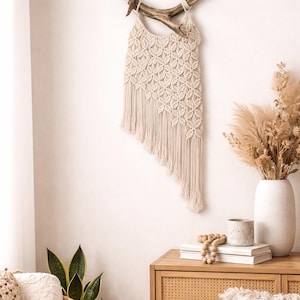 May include: A cream-coloured macrame wall hanging with a fringed bottom is suspended from a piece of driftwood. The handmade textile features a repeating floral pattern. The decor is displayed against a white wall, creating a bohemian aesthetic.