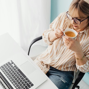 May include: A person seated at a white desk, holding a white coffee cup with a brown liquid. The person is wearing a tan and white striped shirt, blue jeans, and glasses. A laptop is on the desk.
