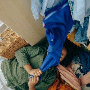 May include: A person surrounded by a pile of colourful clothing, including a green button-down shirt, blue trousers, and other garments. A wicker basket and a wooden drawer are also visible.