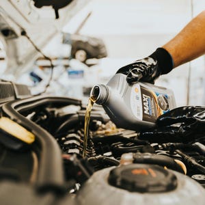 May include: A person wearing black gloves pouring oil from a gray MAXI Performance oil bottle into a car engine. The oil stream is golden, and the engine components are visible.