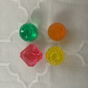 Puede incluir: Cuatro pequeños juguetes redondos, coloridos, parecidos a caramelos. Los juguetes son verde, naranja, rojo y amarillo. Los juguetes están sobre una superficie blanca.