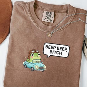 May include: Espresso-colored t-shirt featuring a cartoon frog driving a light blue car, wearing a pilot's cap. A speech bubble says "Beep Beep, Bitch." A silver necklace with a pendant is draped on the shirt.