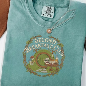 May include: Light green t-shirt with a round graphic featuring a hobbit door, table with food, and the text "Second Breakfast Club." The shirt has a crew neck and short sleeves. A silver necklace with a pendant is on the shirt.
