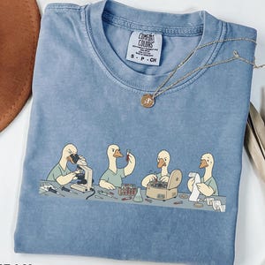 May include: A blue jean colored t-shirt with a graphic print of four geese in a laboratory setting. The geese are wearing scrubs and using scientific equipment like a microscope, test tubes, and a centrifuge. The shirt has a Comfort Colors label.