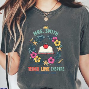 Puede incluir: Una camiseta gris carbón con un estampado gráfico que incluye el texto "MRS. SMITH" sobre un libro abierto coronado por una manzana roja, rodeado por una corona de hojas tropicales, flores de hibisco, estrellas de mar, un lápiz y una regla.