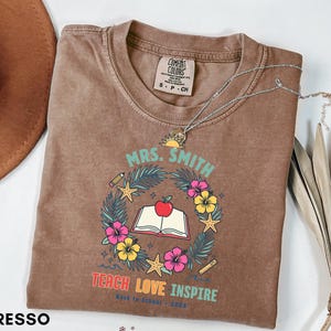 May include: An espresso-colored t-shirt featuring a graphic of an open book with a red apple on top, surrounded by a wreath of tropical leaves, hibiscus flowers, starfish, a pencil, and a ruler. The text reads MRS. SMITH, TEACH LOVE INSPIRE.