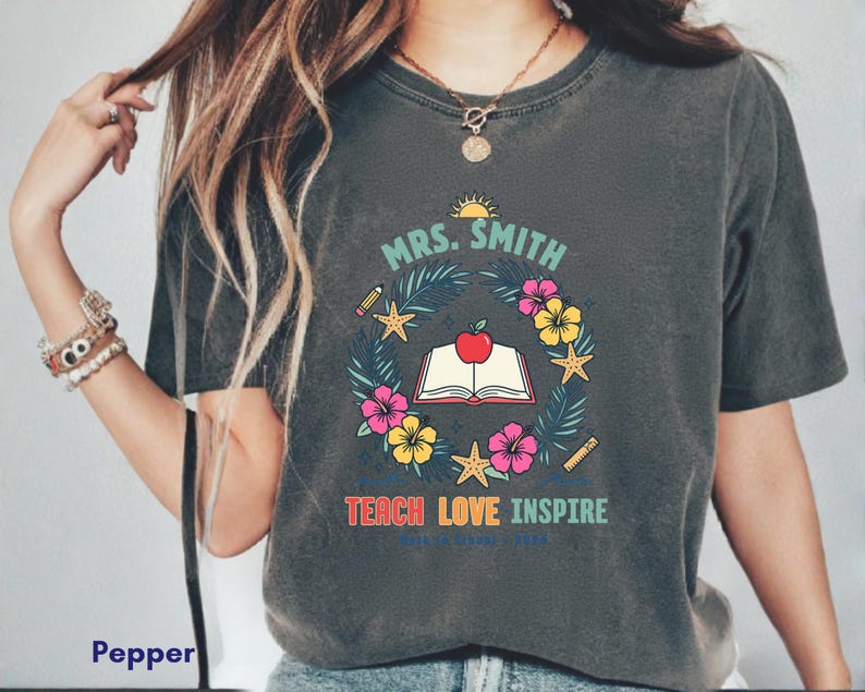 Puede incluir: Una camiseta gris carbón con un estampado gráfico que incluye el texto "MRS. SMITH" sobre un libro abierto coronado por una manzana roja, rodeado por una corona de hojas tropicales, flores de hibisco, estrellas de mar, un lápiz y una regla.